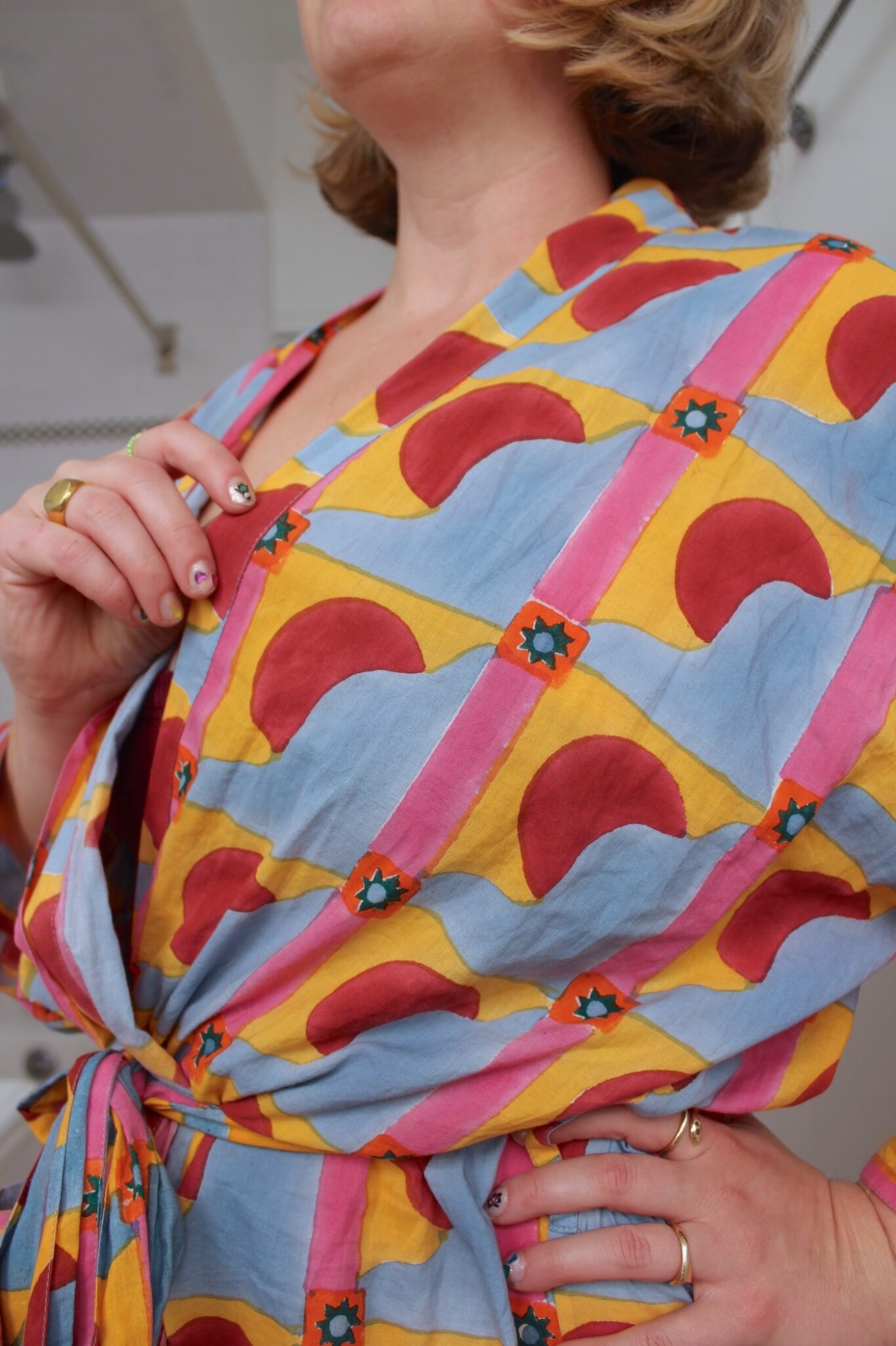Person wearing a colorful patterned wrap dress with red arches, blue stars, and yellow, pink, and blue stripes, hand resting on hip.