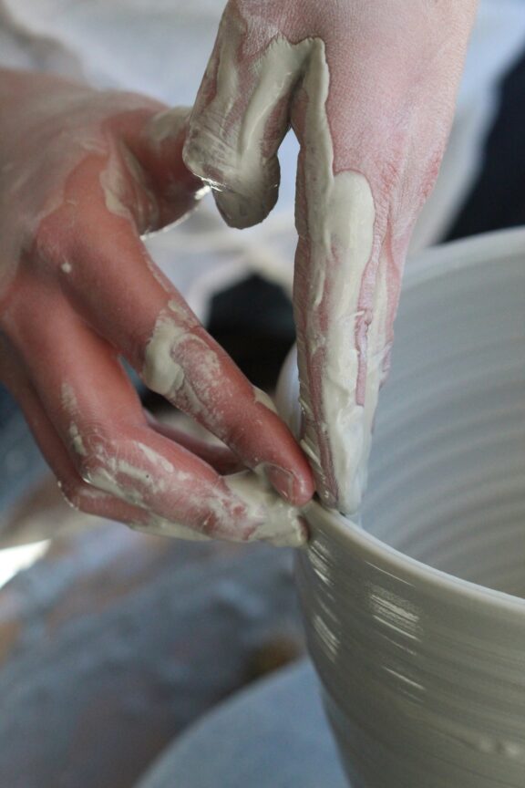 Close-up of hands shaping a clay pot on a pottery wheel, with fingers coated in wet clay.