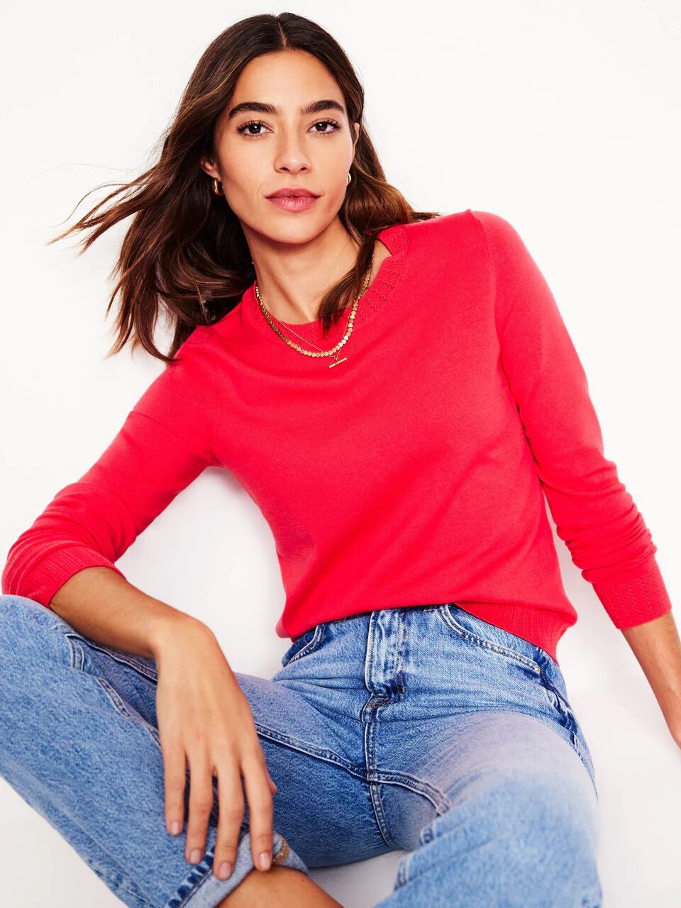 A person wearing a red sweater and blue jeans sits against a white background, looking at the camera.
