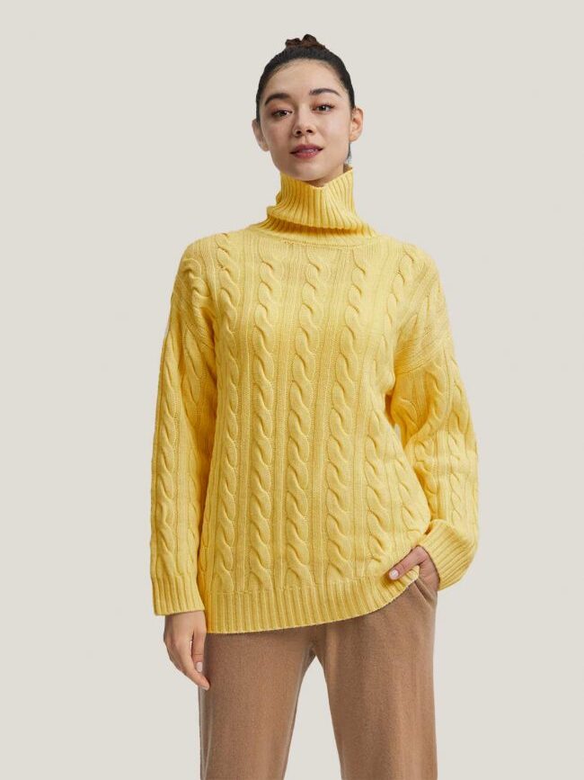 Person wearing a yellow cable-knit turtleneck sweater and beige pants stands against a plain background.