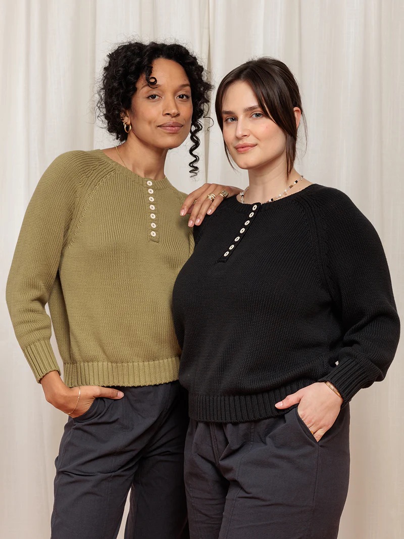 Two people stand closely, wearing modern knit sweaters—one in olive green and the other in black—against a light curtain background. Both have relaxed poses and slight smiles.