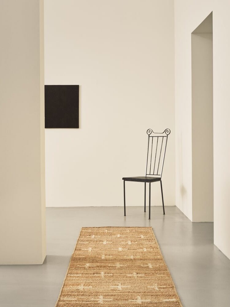 A minimalist room with a woven rug on a gray floor, a black metal chair, a square dark wall art piece, and off-white walls with doorways.