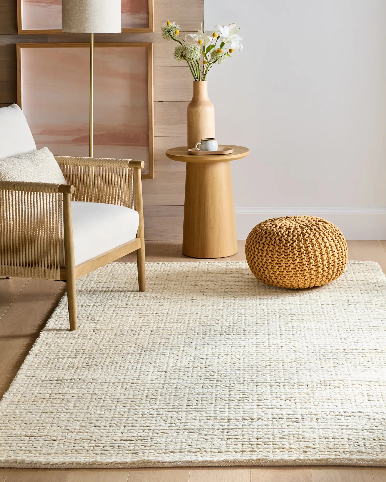A modern living room features a cream woven rug, wooden armchair with white cushion, round knit pouf, small wooden side table, vase with flowers, and abstract wall art.