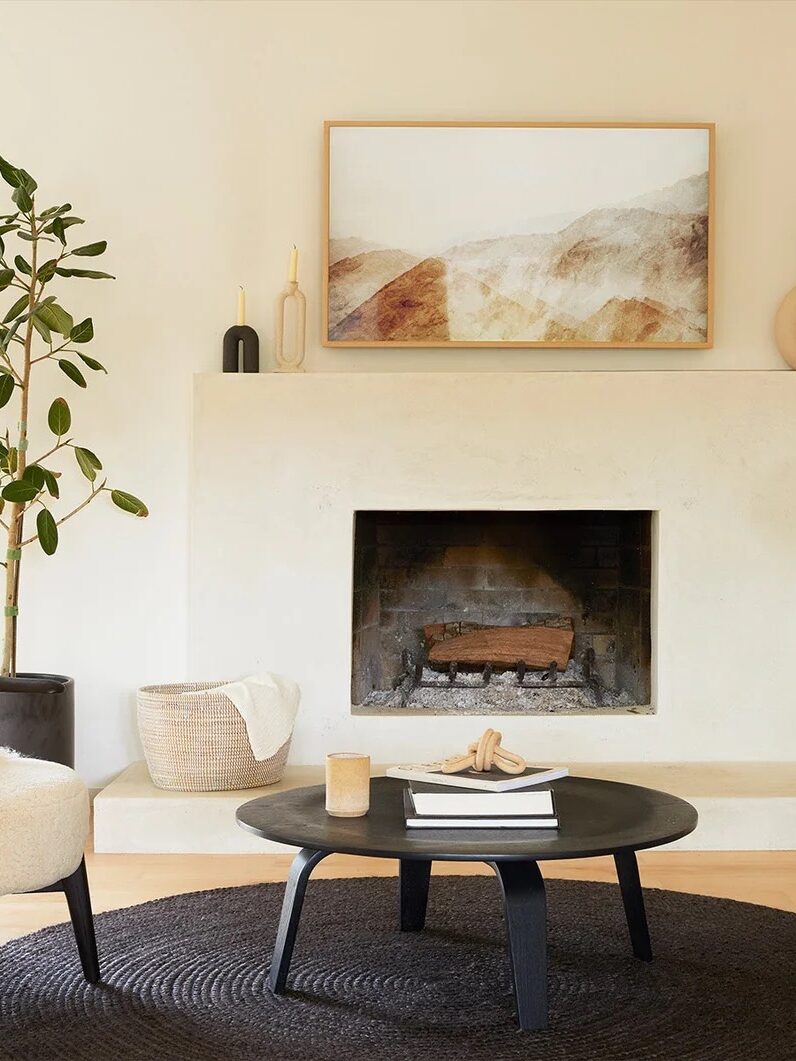 A modern living room with a round black coffee table, a white chair, a bench, a floor lamp, plants, and a fireplace with abstract art above it.