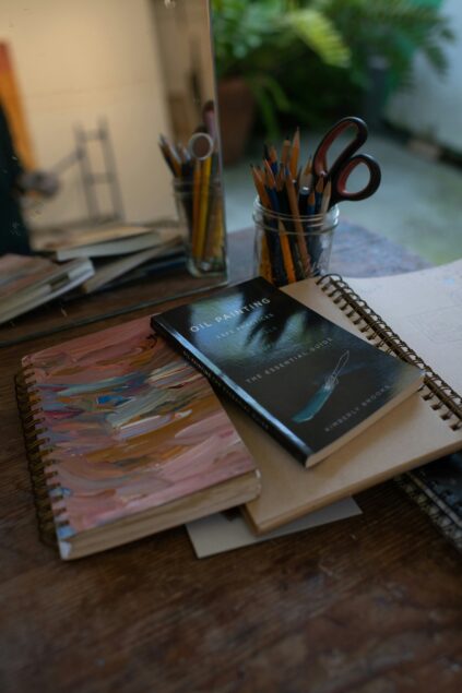 A wooden desk with sketchbooks, a book titled "Oil Painting," and jars holding pencils and paintbrushes, with a mirror and potted plant in the background.