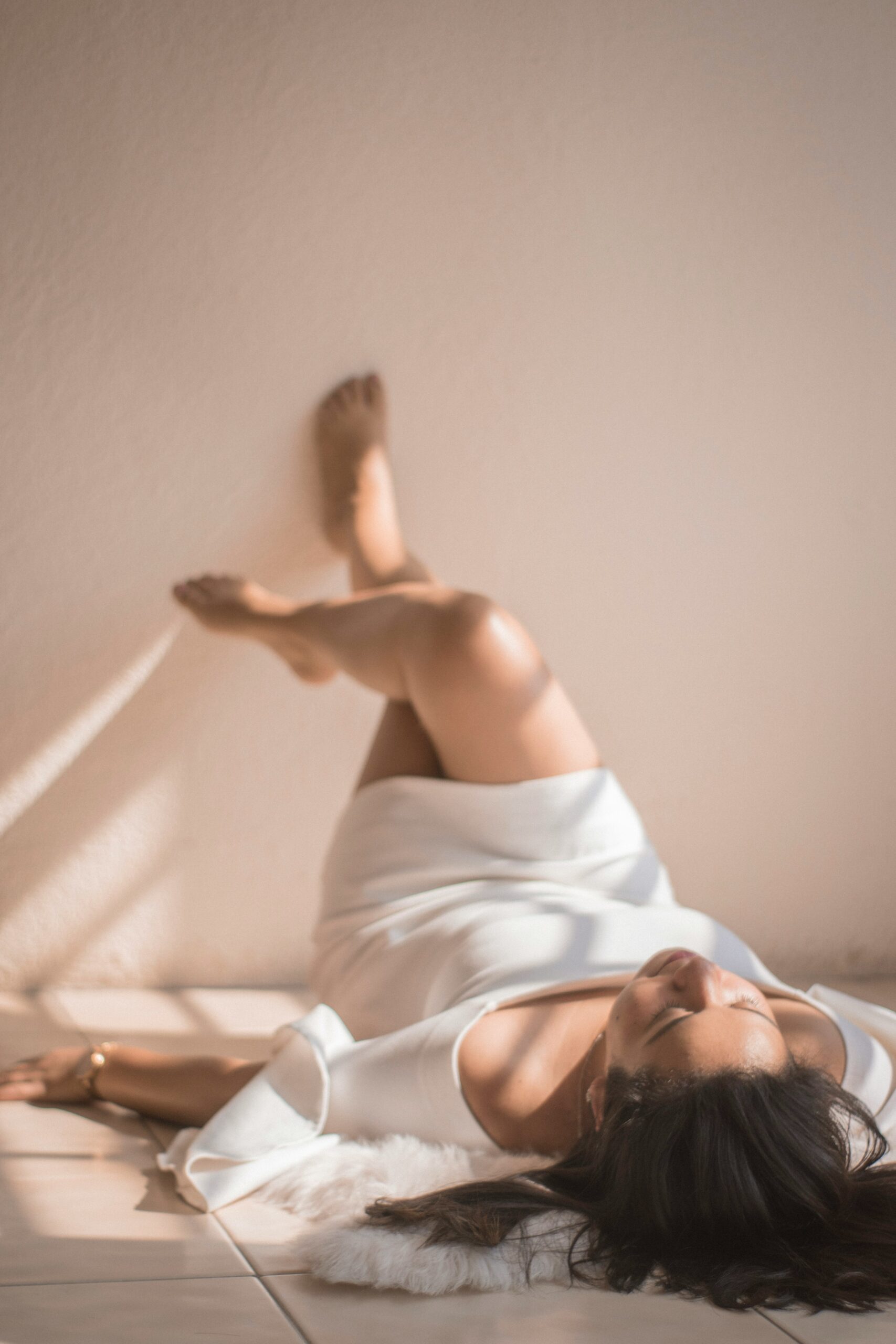 Woman in a white dress lies on the floor with her legs propped up against a light-colored wall, bathed in natural sunlight.