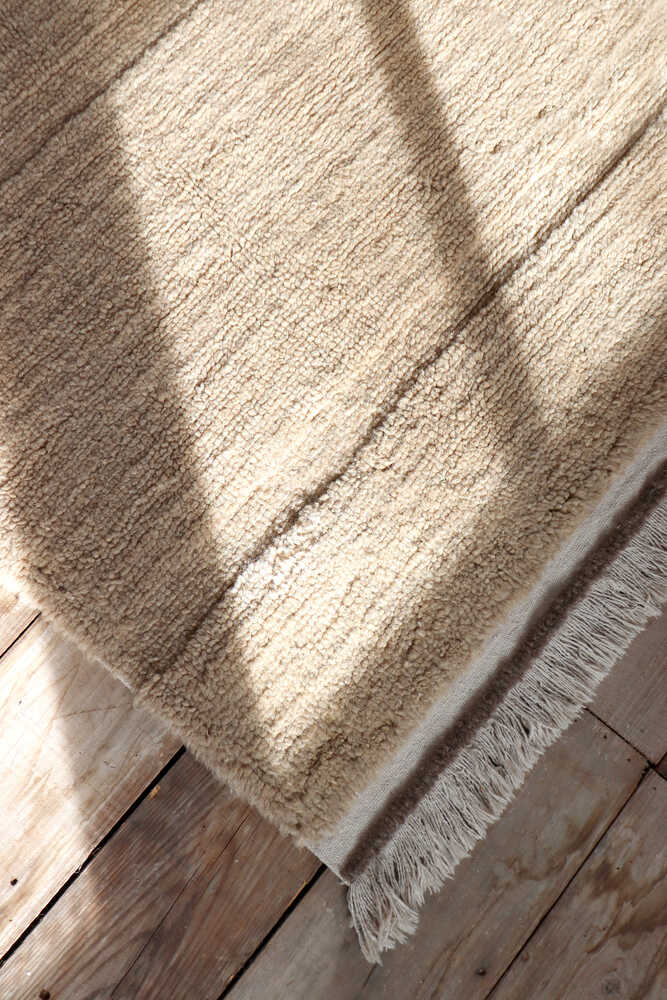 A beige textured rug with fringe on wooden floor, partially illuminated by sunlight and shadow.