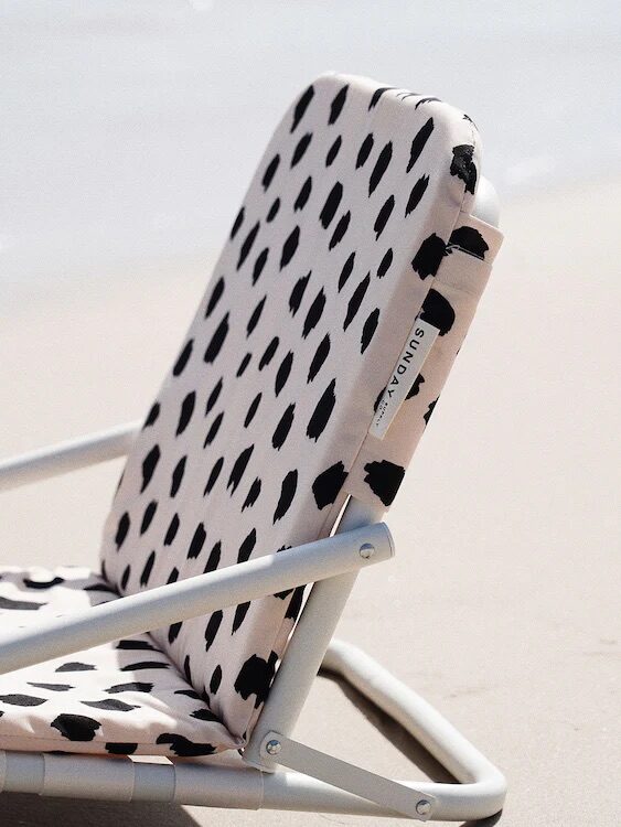 A close-up of a beach chair with a white and black spotted pattern, positioned on sandy ground by the edge of water.