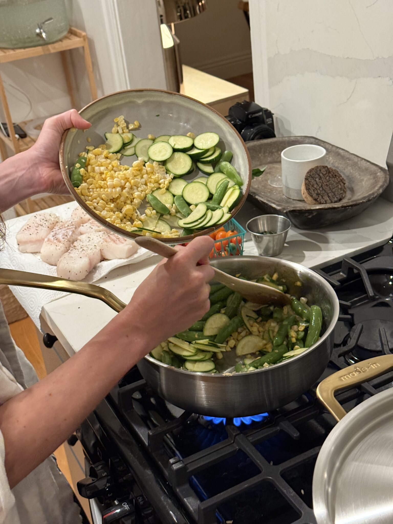 A person adds sliced zucchini, corn, and snap peas from a bowl into a pan on a stove. Raw fish fillets and ingredients are arranged nearby.