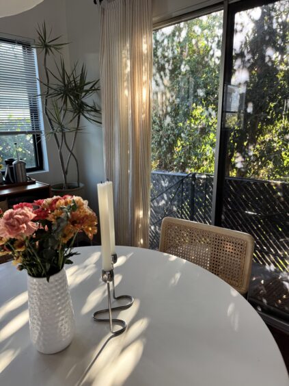 Sunlight filters through curtains onto a round table with a vase of flowers, two tall candles, and a woven chair by a glass door overlooking greenery.