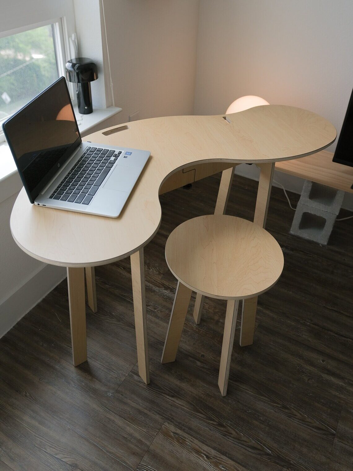 A curved wooden desk with a matching stool holds an open laptop in a bright room. A small lamp and magazine rack are on the wall nearby.