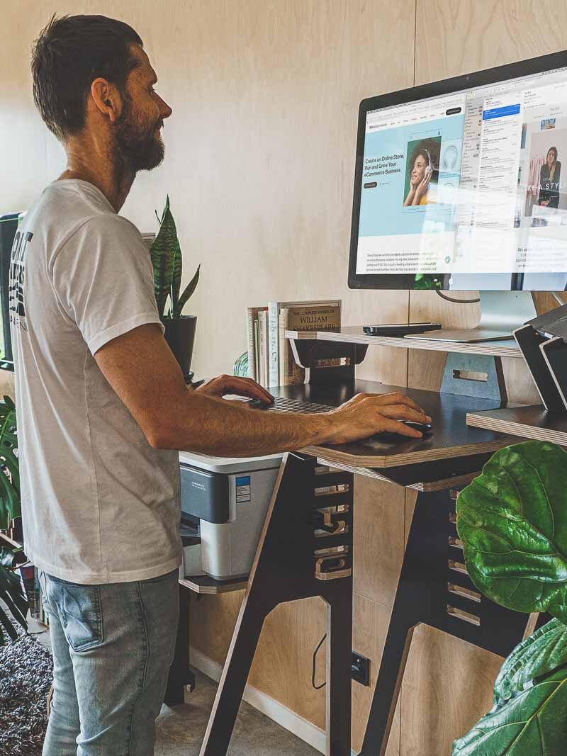 Man using a standing desk with a laptop and monitor in a home office, surrounded by plants and natural light from a window.