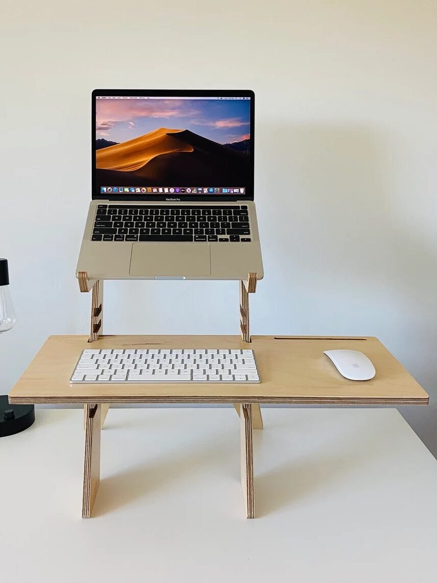 Laptop on a standing wooden desk with a keyboard and mouse below. A minimalist black lamp is on the left side of the desk.