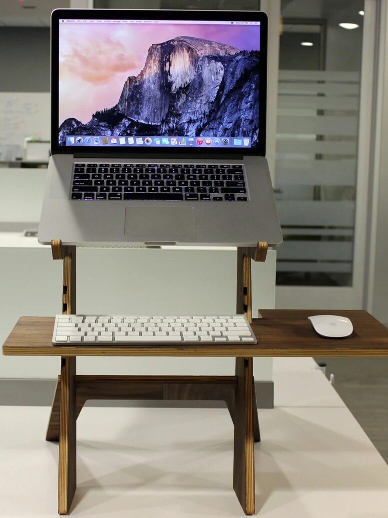 A laptop on a wooden stand with a keyboard and mouse attached. The background shows an office setting with glass doors and a whiteboard.