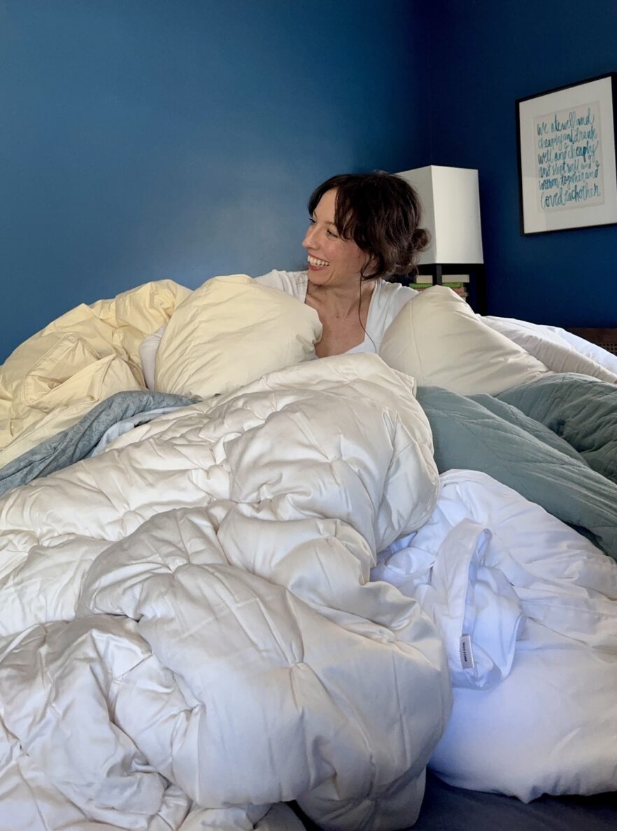 A woman sits on a bed with an organic mattress, surrounded by a large pile of comforters and pillows, smiling. The room has blue walls and framed art on the wall.