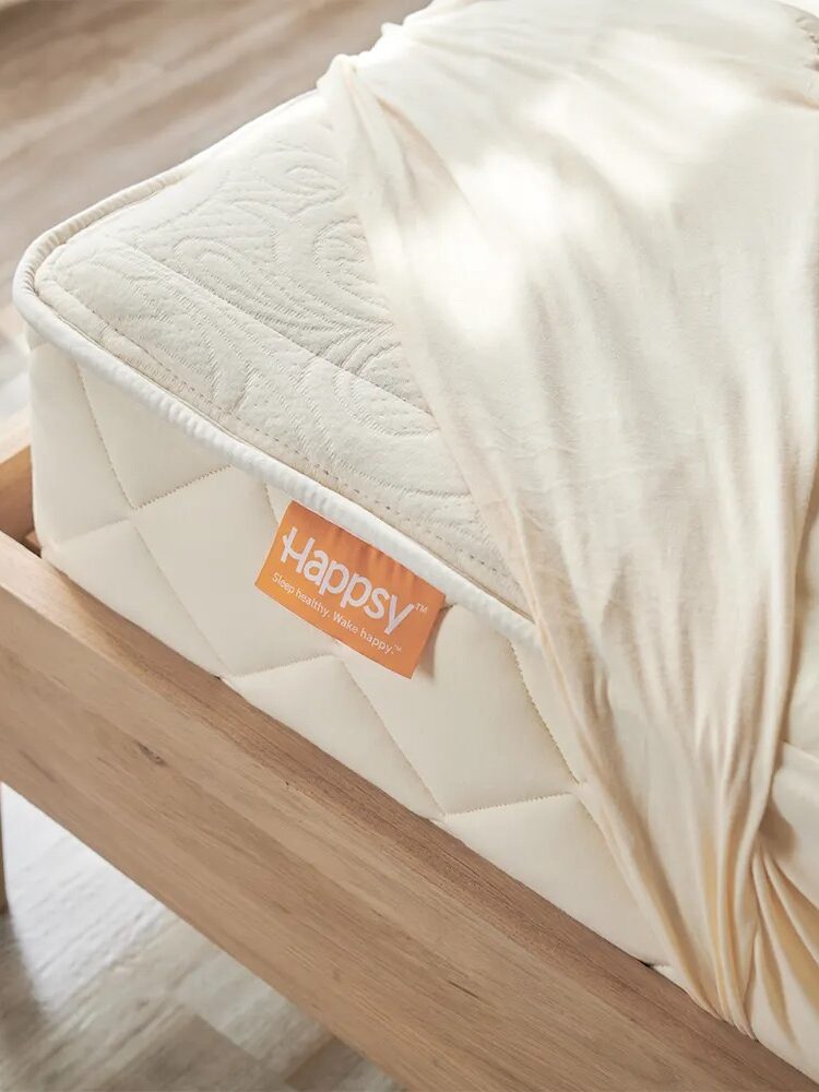 A wooden bed with a neatly placed mattress. A corner of the cream-colored cover is pulled back, revealing a label with the brand name "Happsy." Natural light illuminates the scene.