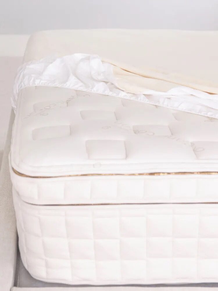 A close-up of a white mattress partially covered with a fitted sheet. The mattress has a quilted pattern and an exposed top corner.