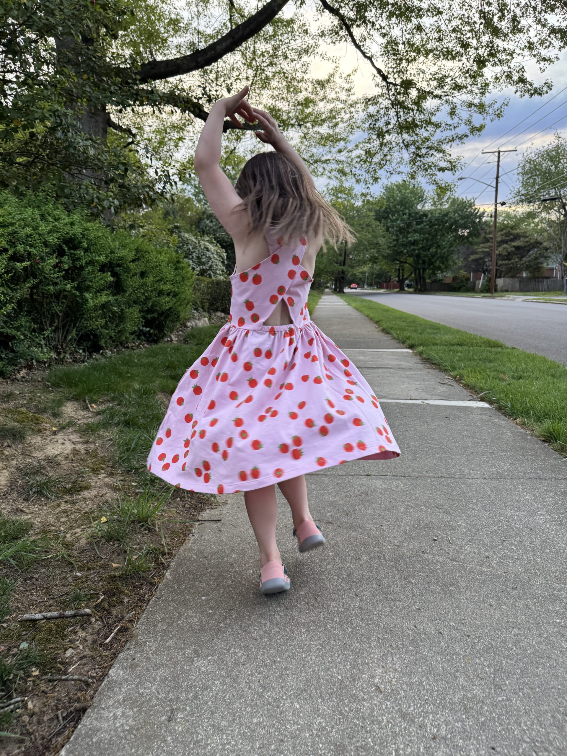 A young girl in a pink dress with red dots spins on a sidewalk near grass and trees on a cloudy day.
