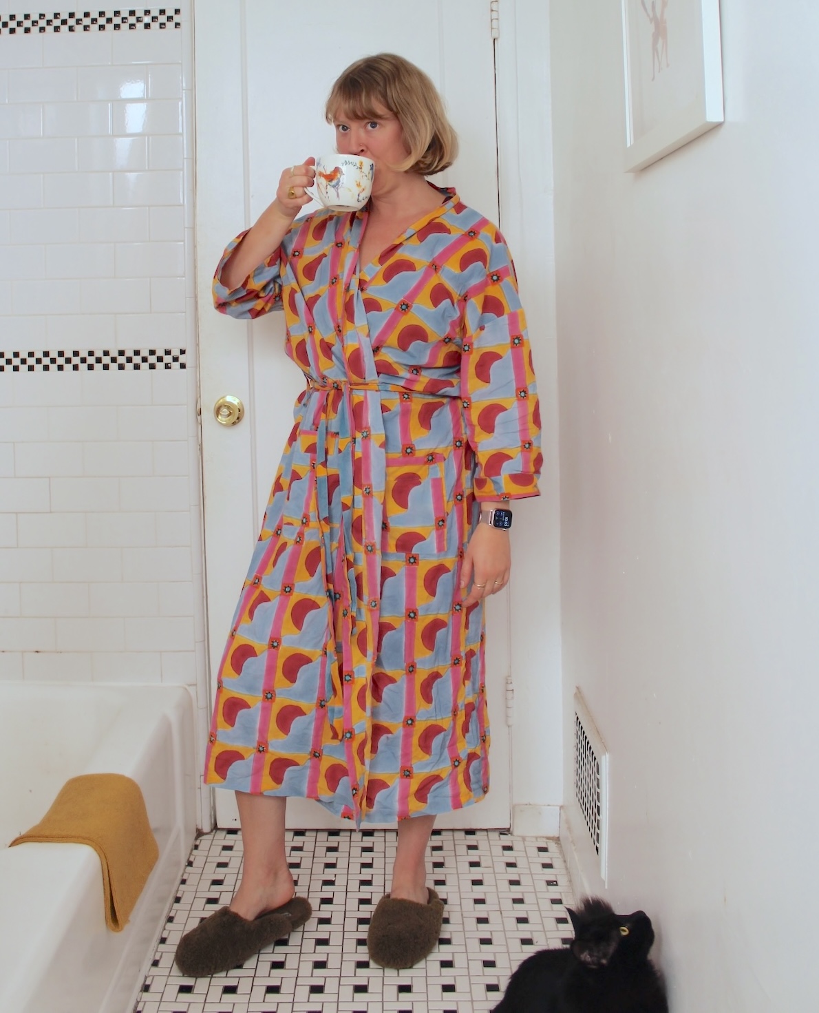 A person in a colorful bathrobe drinks from a mug in a bathroom with white tile. A black cat sits on the floor near their feet.