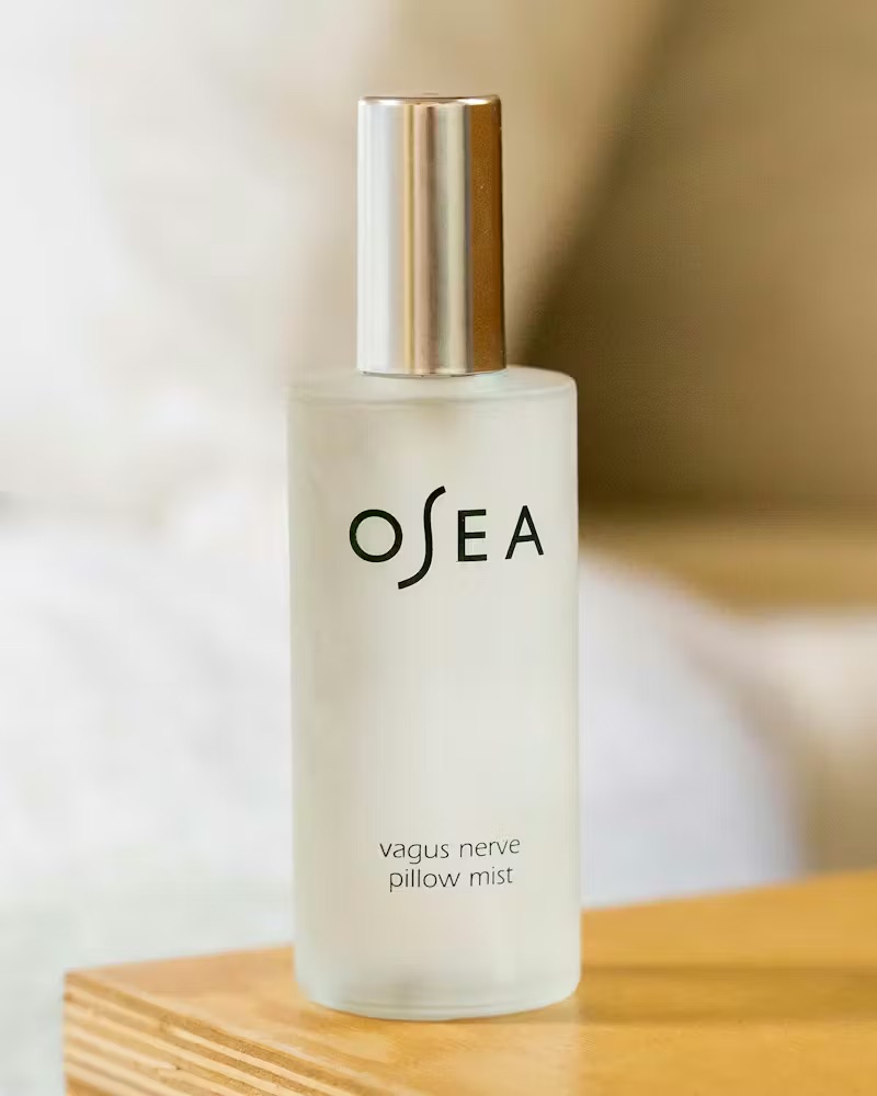 A frosted spray bottle labeled "OSEA vagus nerve pillow mist" sits on a wooden surface, with a blurry background.