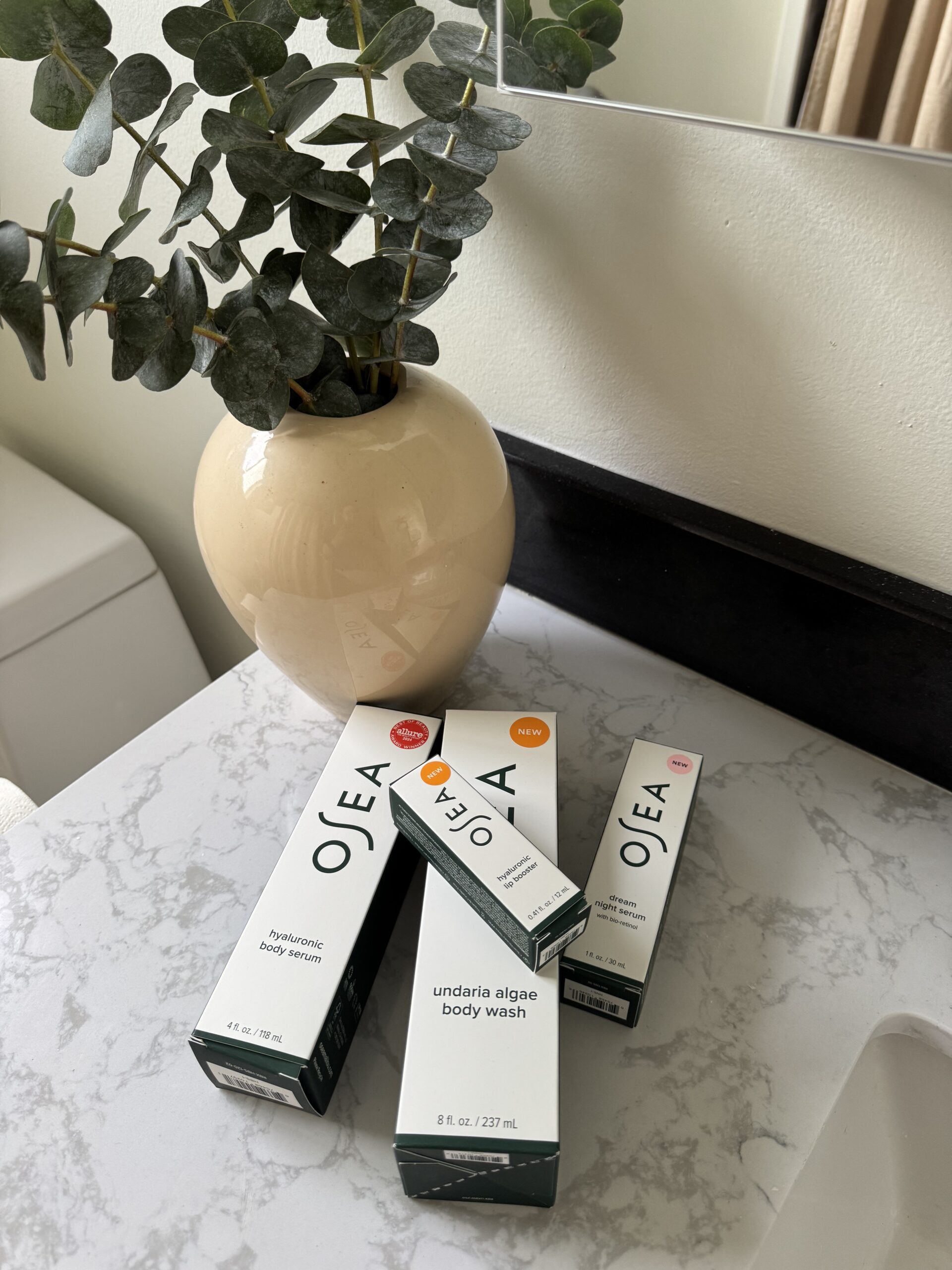 Three OSEA skincare product boxes are arranged on a white marble bathroom counter next to a beige vase with green eucalyptus branches.