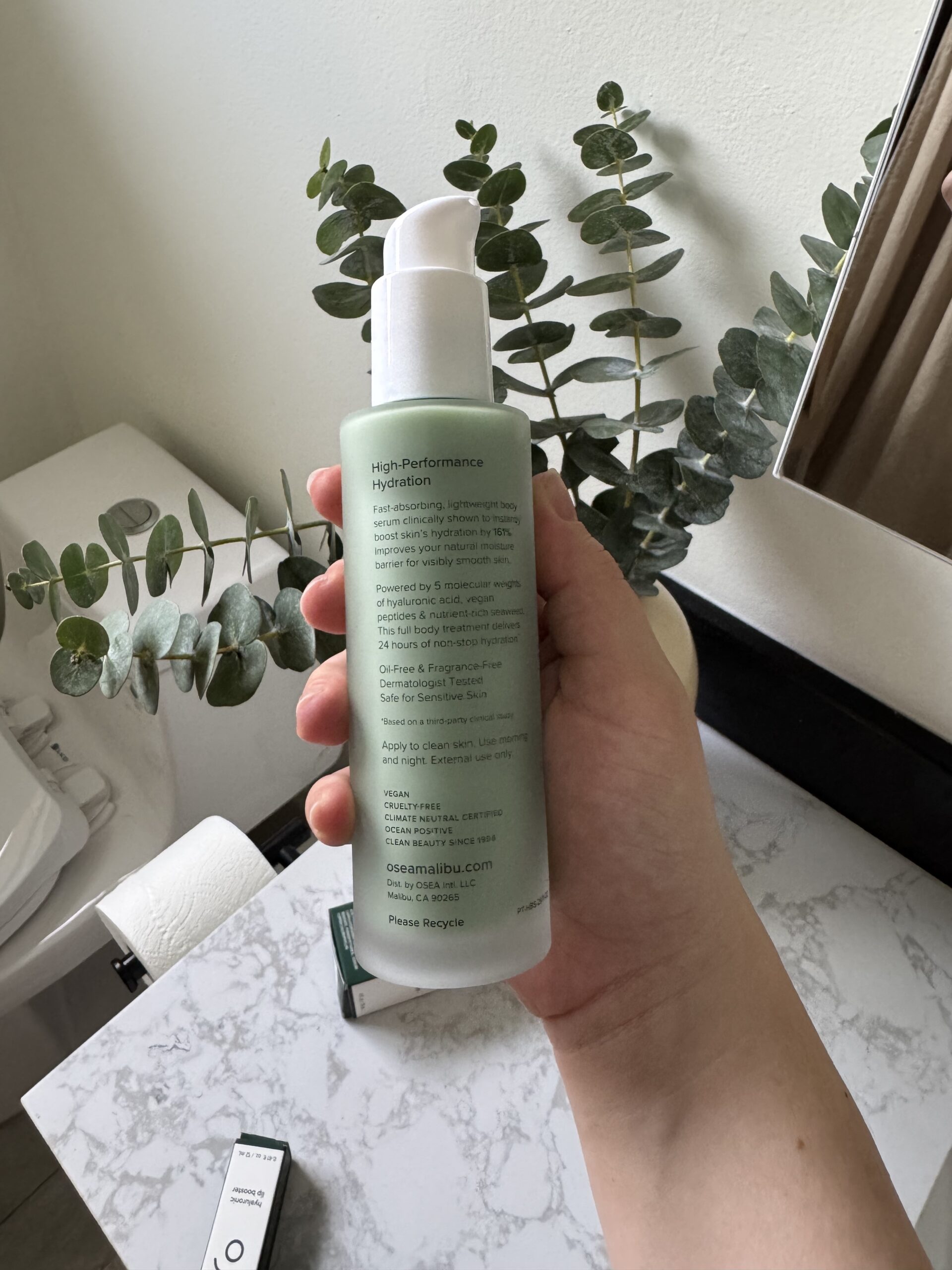 A hand holds a green bottle of Osea Malibu High Performance Hydration lotion in a bathroom with eucalyptus branches in the background.