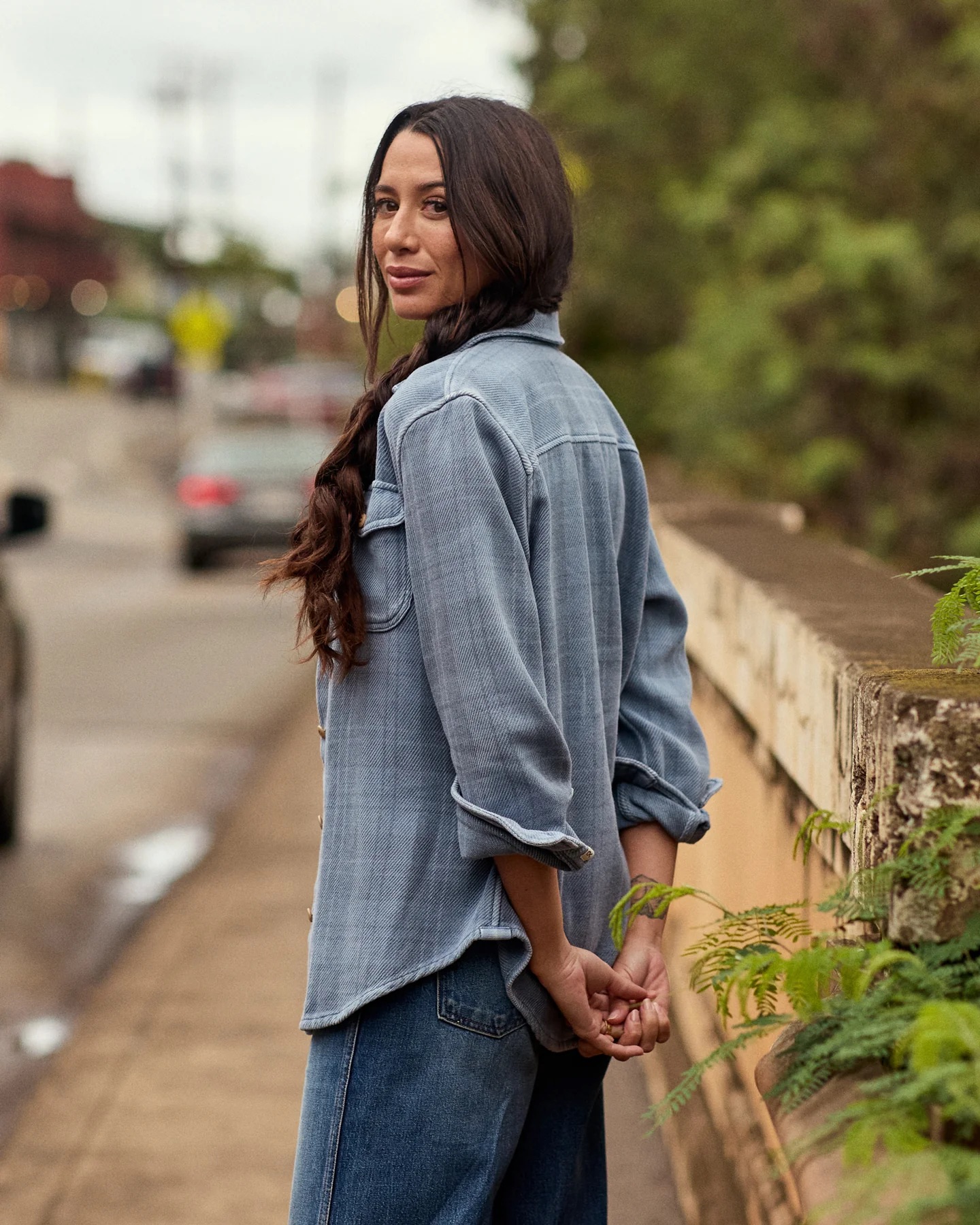 Woman with long dark hair in a braid stands on a sidewalk, wearing a blue button-up shirt and jeans, looking over her shoulder toward the camera.