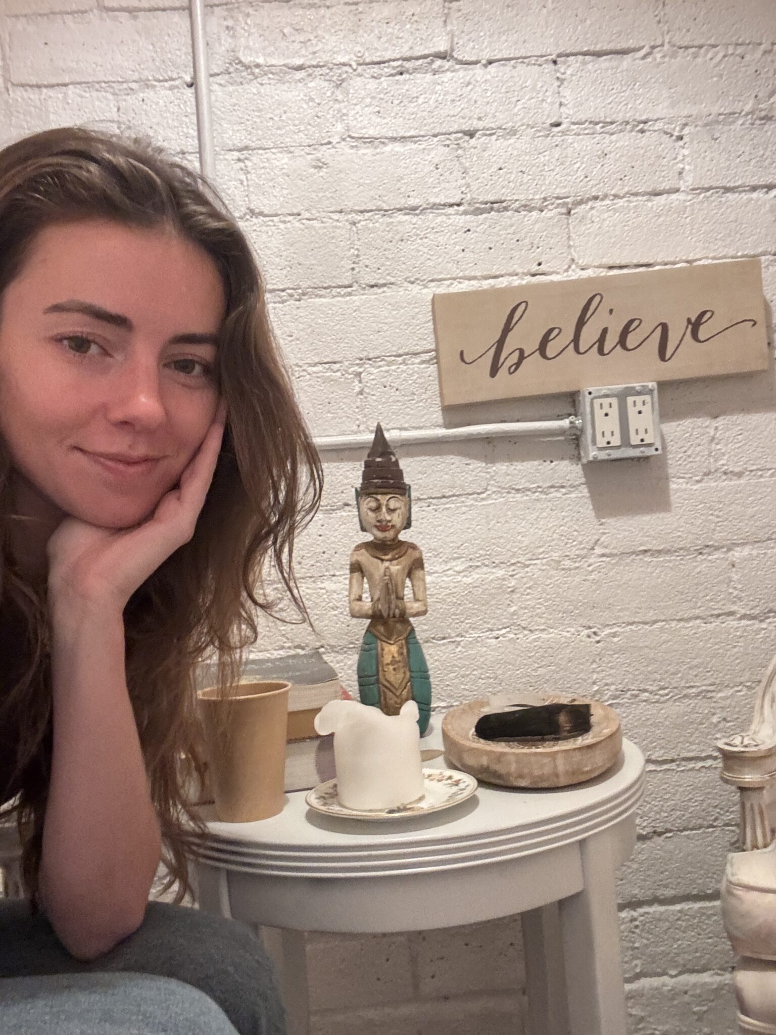 A woman sits beside a small round table with a Buddha statue, candle, wooden bowl, and a sign on the wall that says "believe.