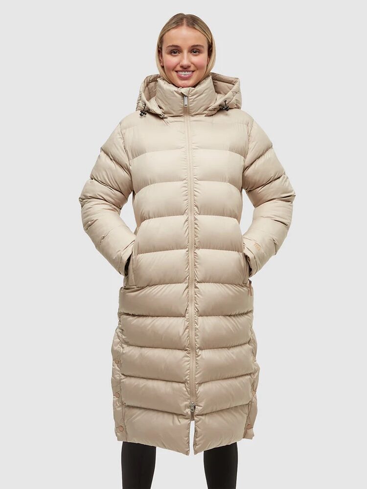 A person stands facing the camera wearing a long, light beige puffer coat with a hood against a plain background.