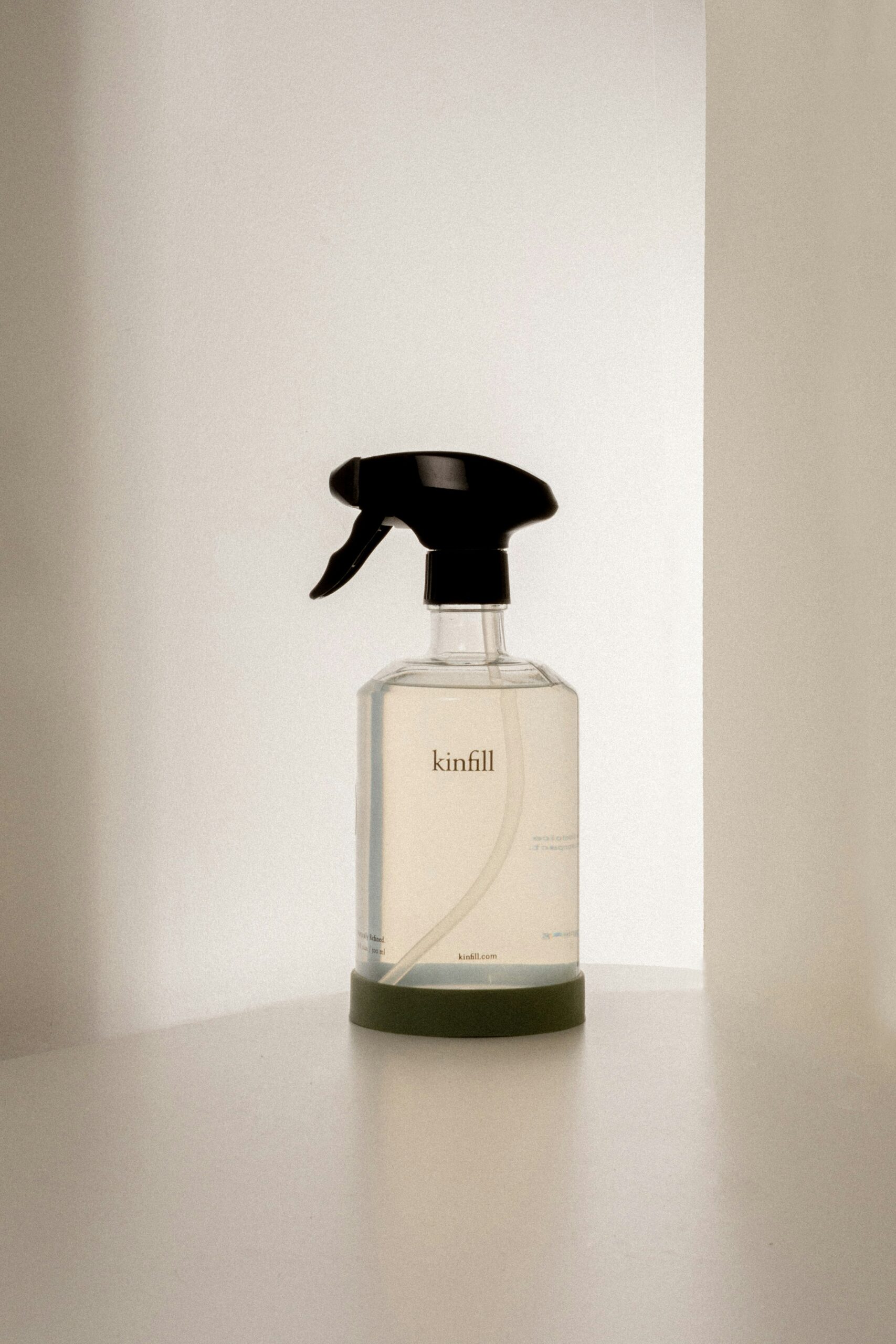A clear spray bottle labeled "kinfill" with a black nozzle, placed on a white surface against a softly lit background.