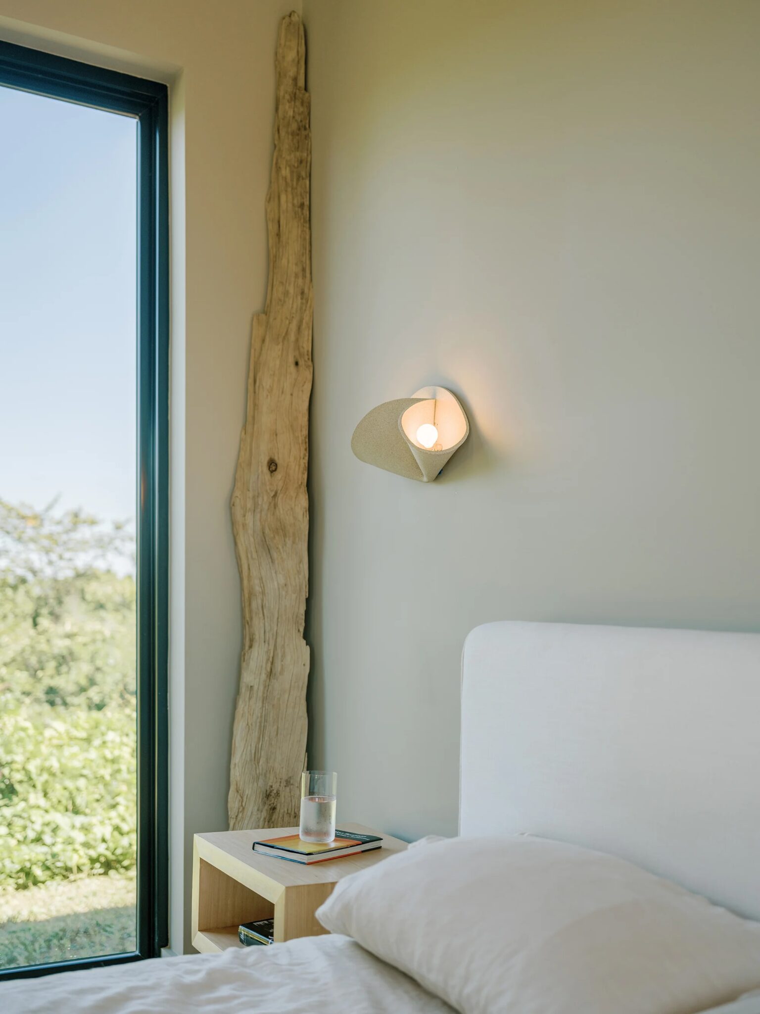 Minimalist bedroom with a modern wall sconce, a wooden nightstand holding books and a glass of water, large window, and rustic wooden decor piece against a pale wall.