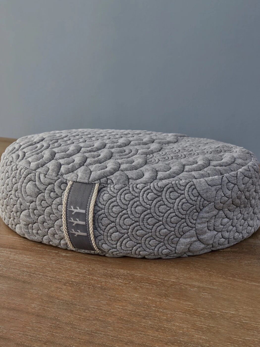 Round gray cushion with a quilted pattern, featuring a fabric loop with decorative stitching, placed on a wooden surface against a gray background.