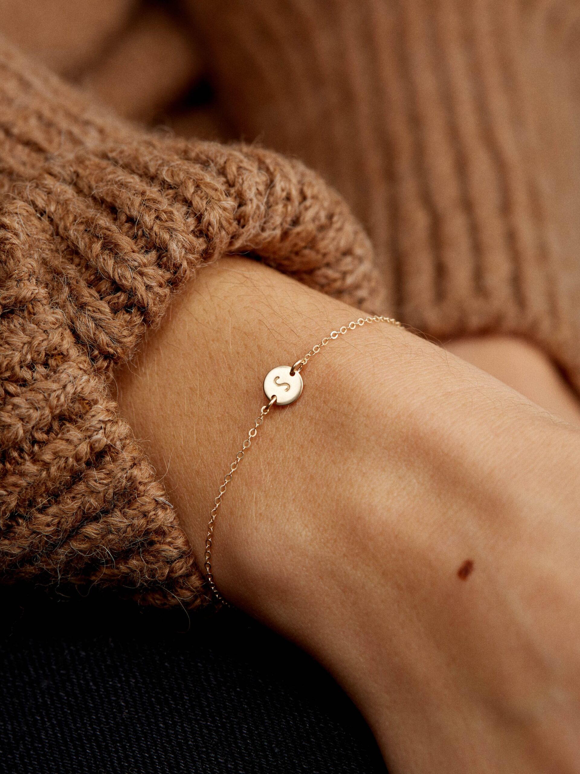 A gold chain bracelet with a round, engraved "S" charm is worn on a wrist in a brown knit sweater.