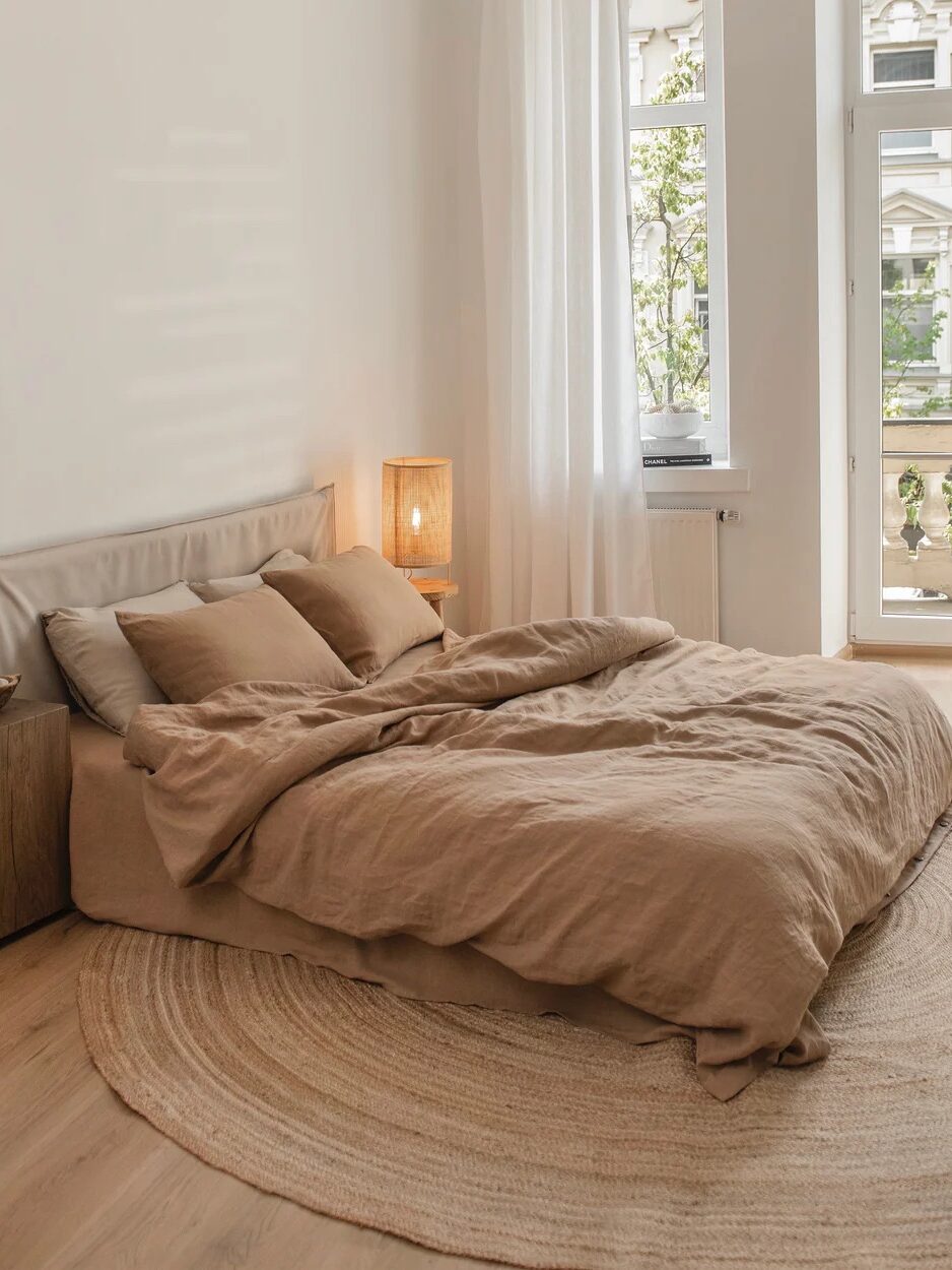 Minimalist bedroom with a low bed covered in beige linens, a circular woven rug, a wooden nightstand with a lamp, and a large window with sheer curtains.