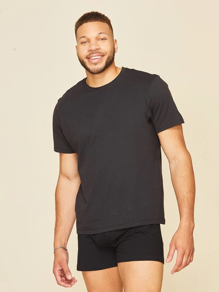 A man stands smiling, wearing a plain black t-shirt and black boxer briefs against a beige background.