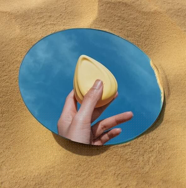 A hand holding a yellow makeup sponge is reflected in a round mirror placed on sand, with blue sky visible in the reflection.
