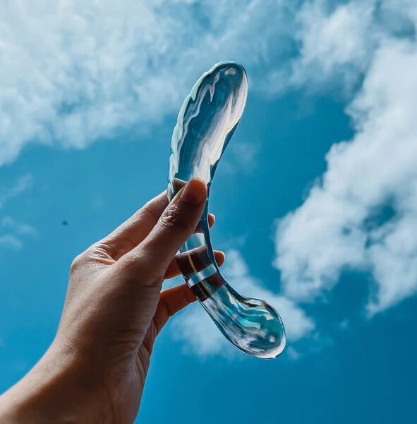 A hand holds a transparent, curved glass object against a blue sky with some white clouds.