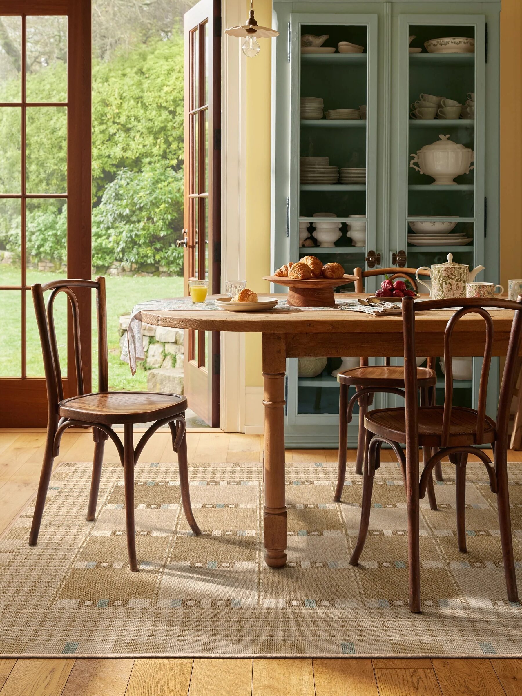 A wooden dining table with chairs is set for breakfast with croissants, fruit, and drinks. A blue cabinet with dishes and large windows are in the background.