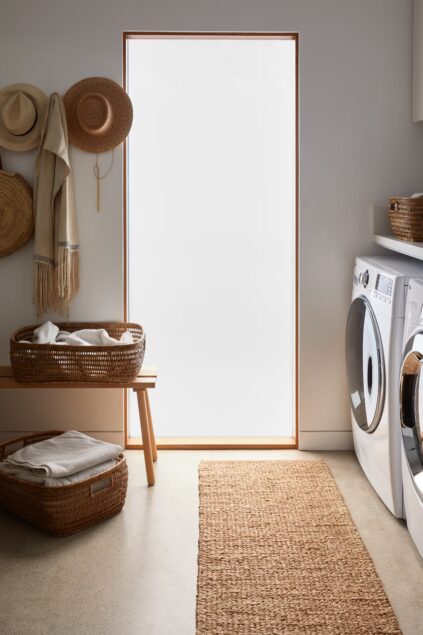 A laundry room with a washing machine, dryer, woven baskets, hats on hooks, a bench, and a textured rug, with light coming through a frosted door.