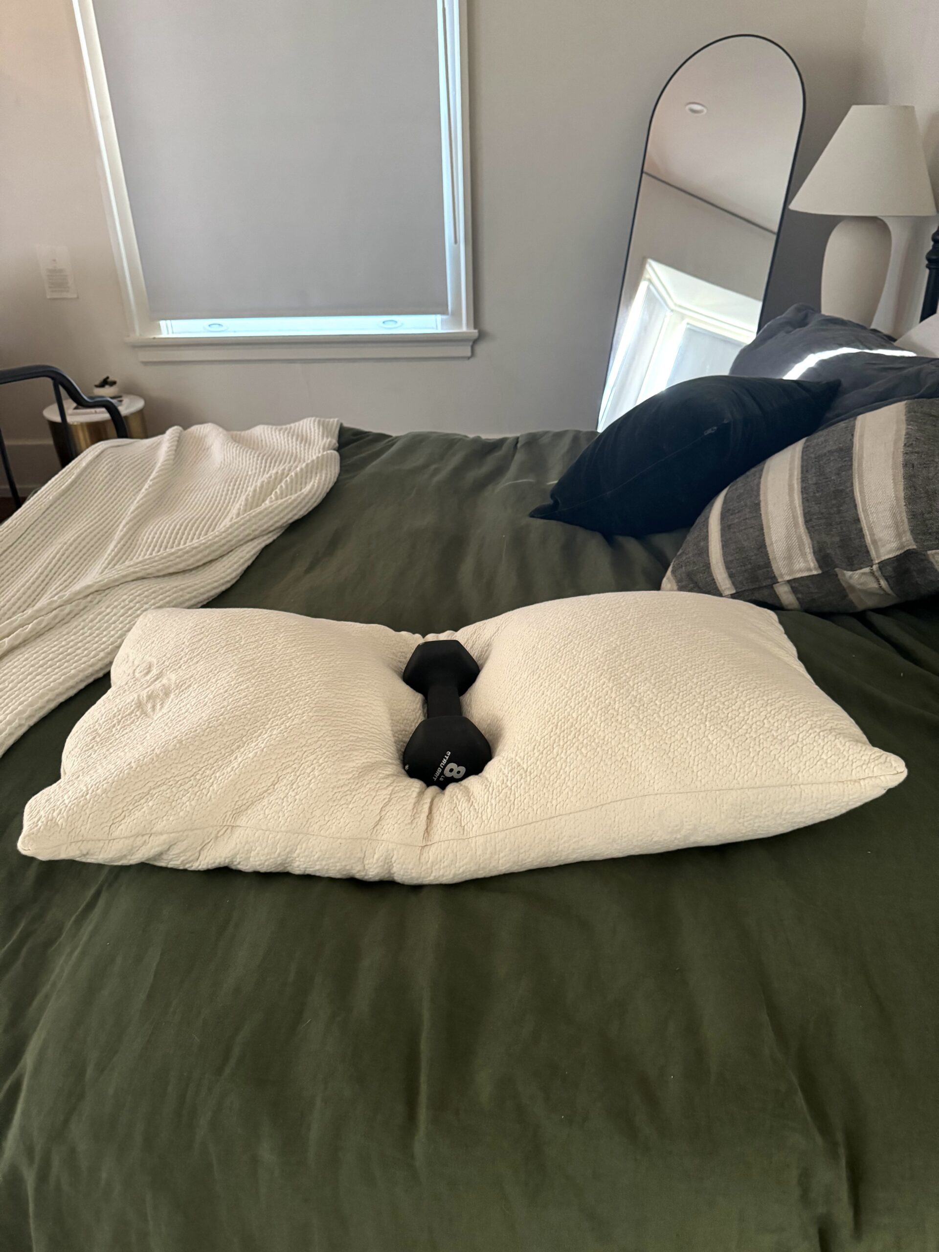 A pillow on a neatly made bed is folded around a black dumbbell. A mirror and lamp are in the background.