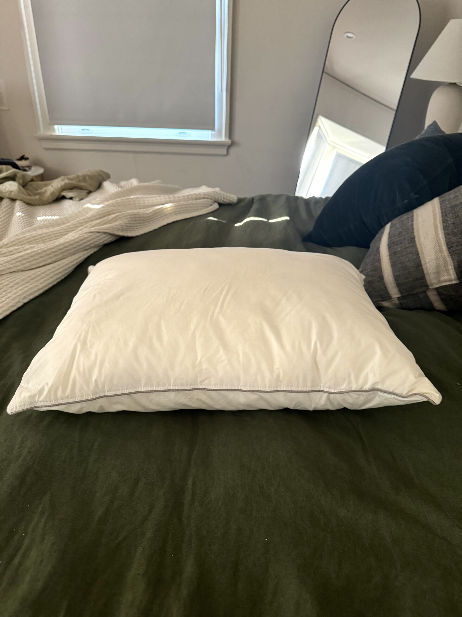 A white pillow is placed on a green bedspread. Behind the bed, a window with a closed shade and a mirror reflecting part of the room are visible. Another pillow and a blanket are on the bed.