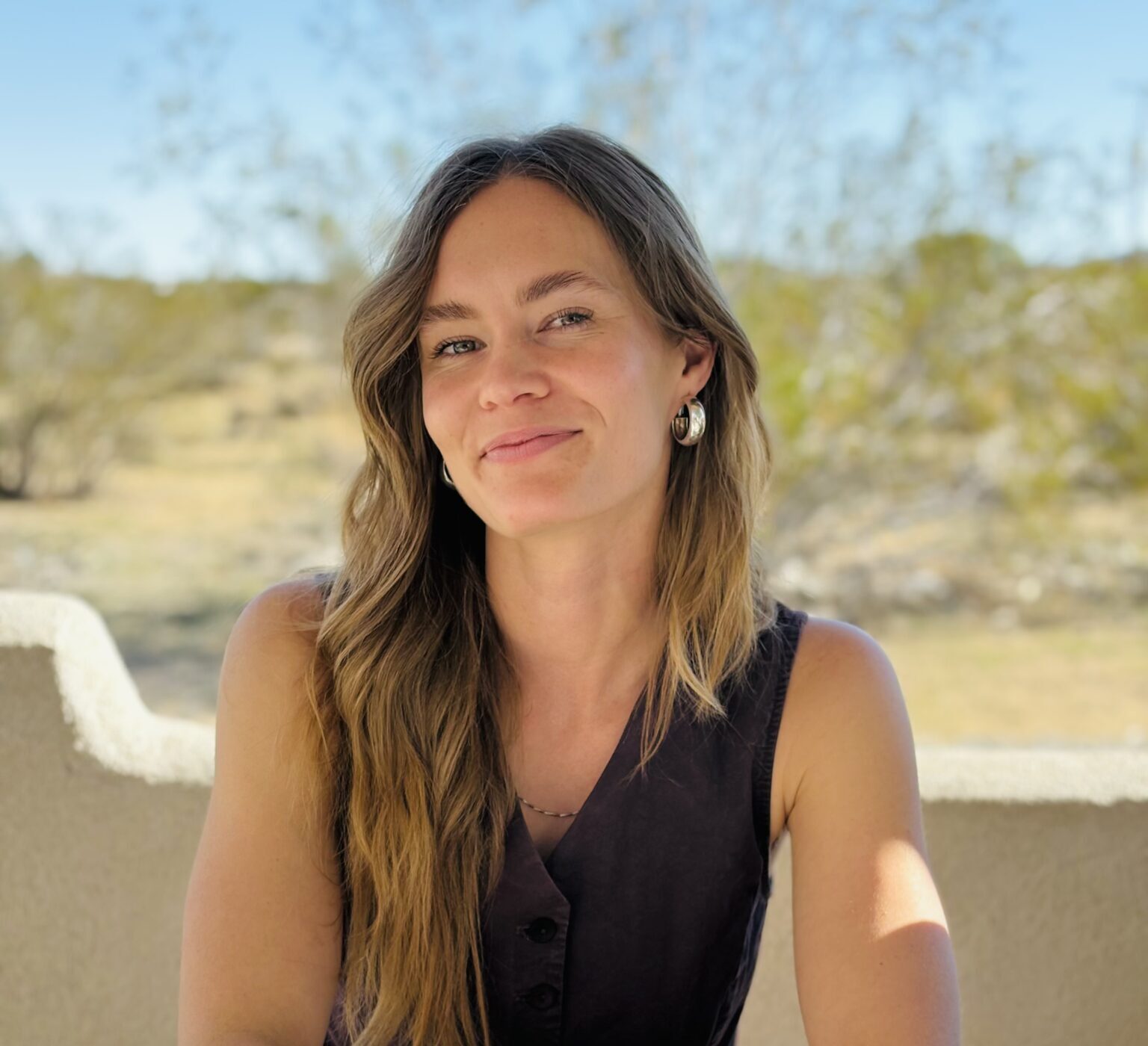 A woman with long brown hair sits at an outdoor table holding a pen, with a notebook, coffee cup, and art supplies in front of her. Desert scenery is visible in the background.