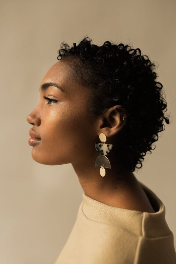 Person with short, curly hair wearing abstract, geometric earrings in profile view against a neutral background.