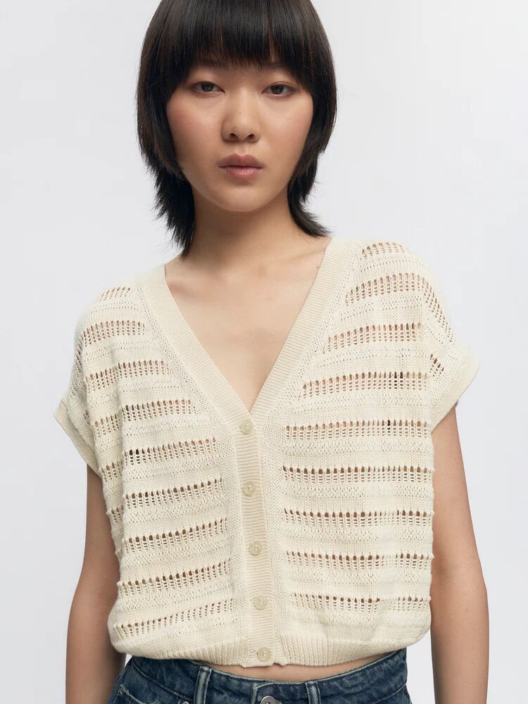 A person wearing a cream-colored, short-sleeve, open-knit cardigan with buttons and blue jeans, standing against a plain white background.