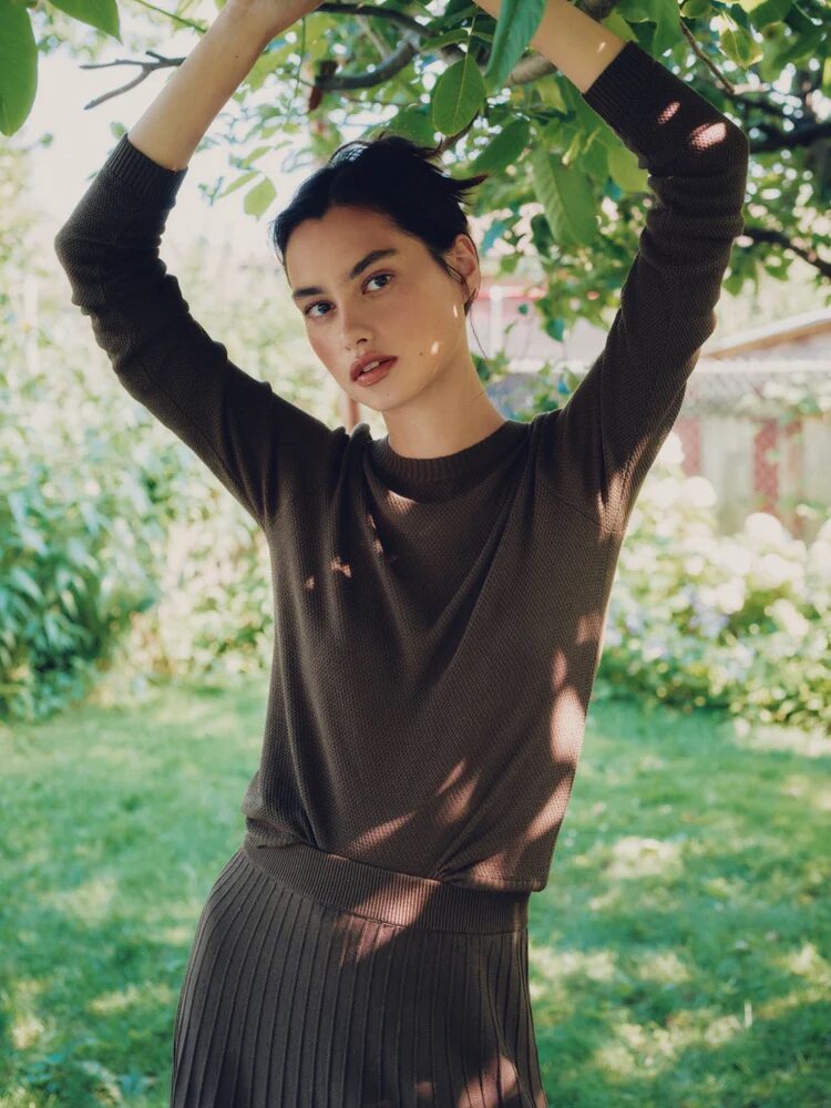 A person in a brown long-sleeve top and pleated skirt stands outdoors under a leafy tree, arms raised, with greenery and a building in the background.