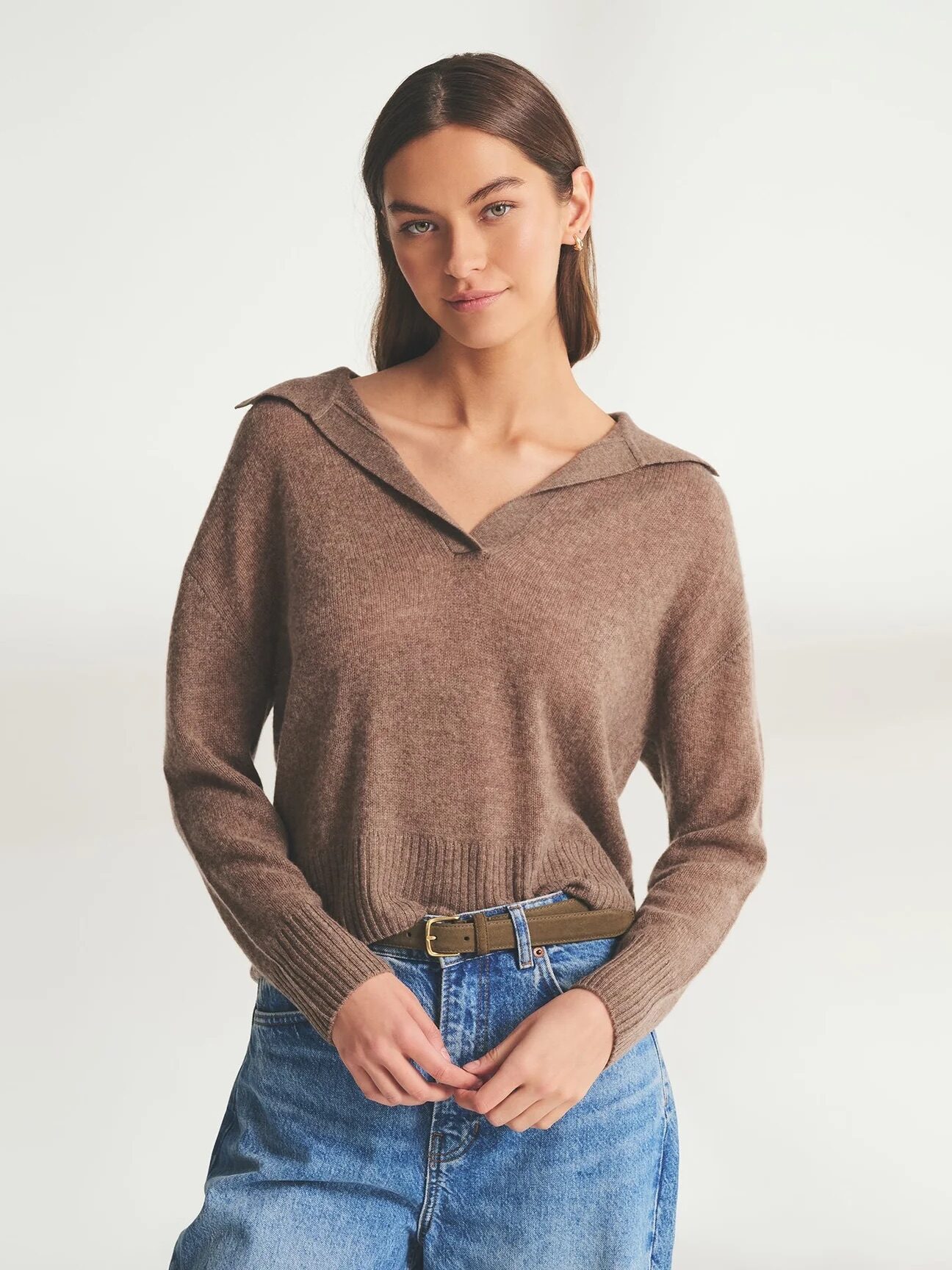 A woman wearing a brown long-sleeve knit sweater with a collar and blue jeans stands against a plain light background.
