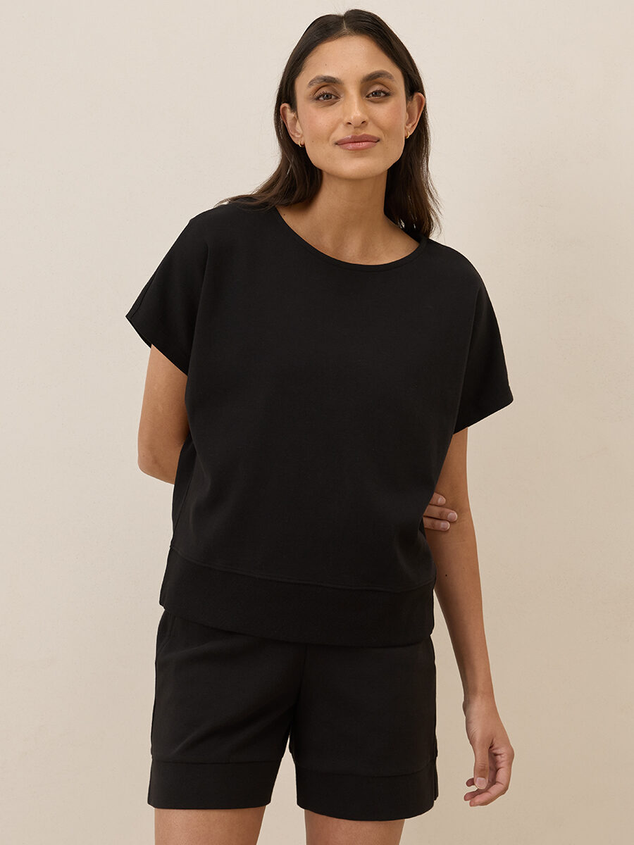 A woman stands against a plain light background wearing a black short-sleeve top and matching black shorts, with one hand behind her back.