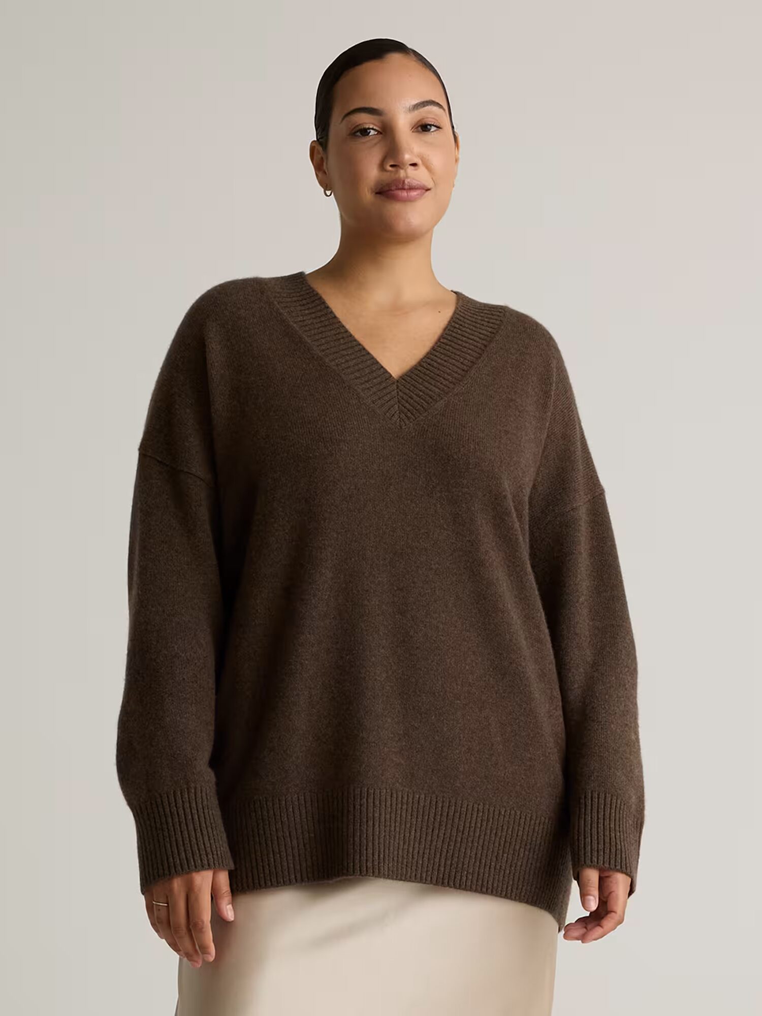 A person wearing a dark brown V-neck sweater and a light-colored skirt stands against a plain, light background.