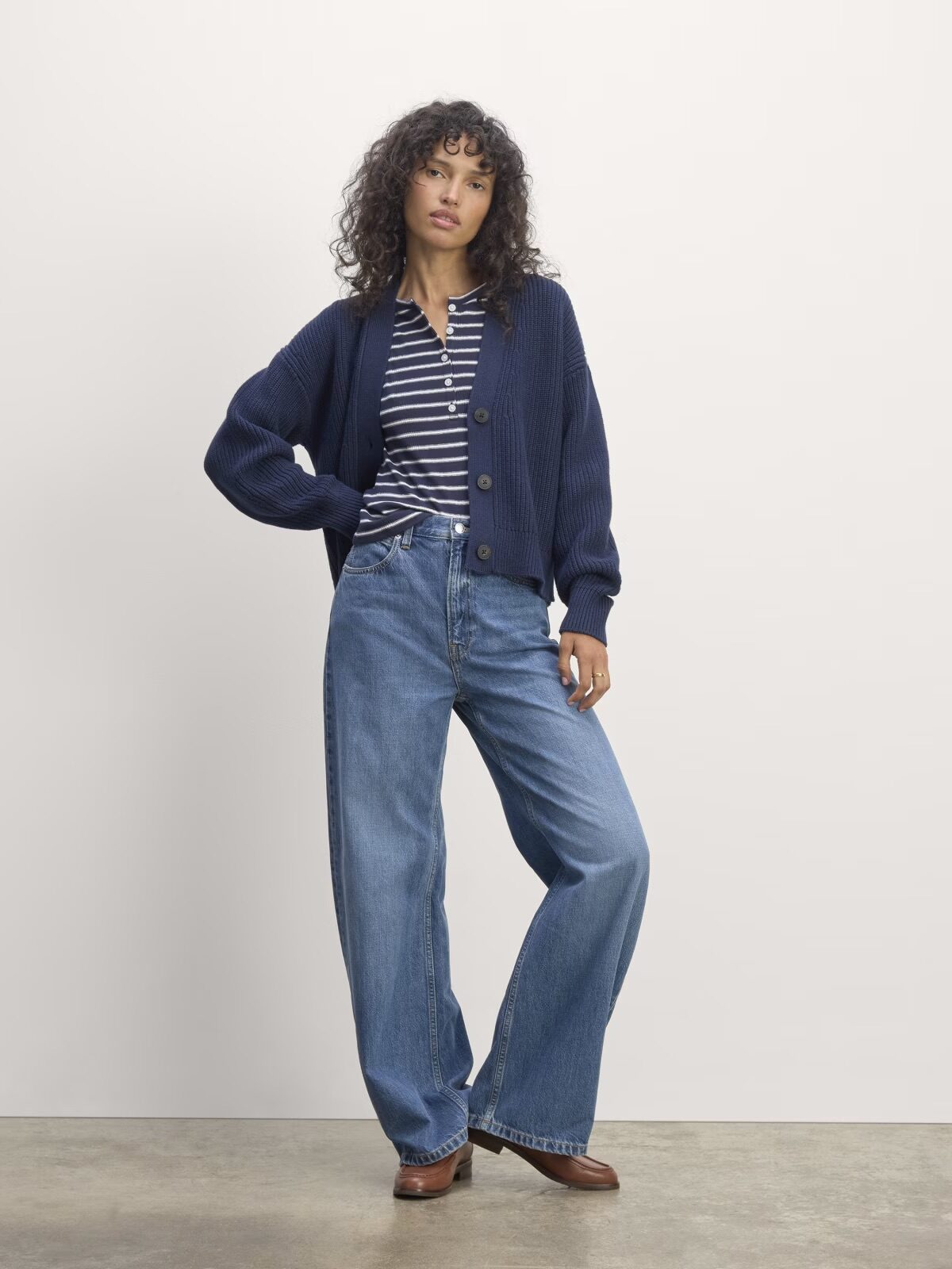 A person stands against a plain background wearing a navy cardigan, striped shirt, wide-leg jeans, and brown shoes.
