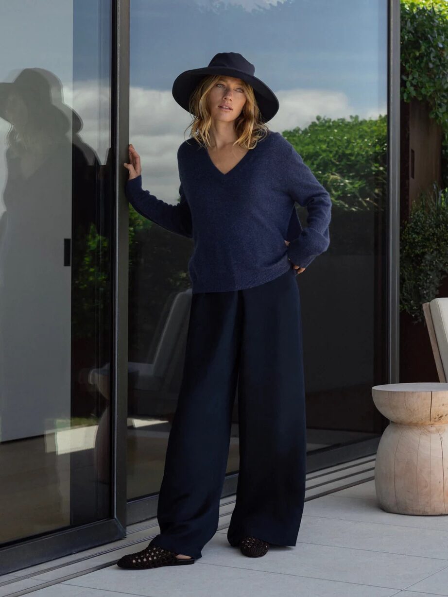 A person wearing a wide-brim hat, navy sweater, and wide-leg pants stands by a glass door on a modern patio with greenery and wooden stools visible.
